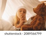 Small photo of Sweet little girl with a teddy bear hold finger on lips showing silence to keep a secret while lies on her bed in a in tent with nightcap.