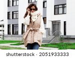 Fashionable woman dressed beige quilted gilet with belt, warm white knitted sweater, scarf, jean. Trend women's clothing for autumn or spring