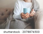 Close up of hands holding blue cup of tea or coffee. Model in white sweater and cozy plaid is sitting, relax at home on sofa. Depth of field, empty space on blurry beige background