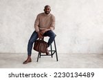 Stylish man sitting on chair and looking camera. Confident fashion model in fashionable outfit turtleneck knitted sweater, plaid trousers, leather shoes and bag. Office clothes style for businessmen