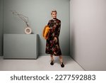 Small photo of Senior fashion female model standing full length in studio wears black maxi floral dress and socks with shoes. Mature woman with short hair