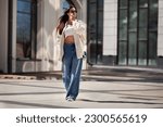 Fashionable woman wearing trendy white bomber jacket, CROP TOP, wide jeans walks on urban city street. Trends clothing of spring and summer.