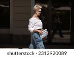Small photo of Sophisticated woman with short hair in white shirt and cargo jeans pants, fashionable accessories earrings, stylish glasses and clutch walking outdoors in city. Close up Fashion style.
