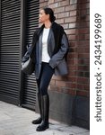 Small photo of Stylish fashion woman Standing the street in multilayer clothes oversized blazer, black leggings, and knee high rubber boots, large bag. Fashionable outfit, street style.