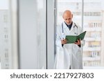 Small photo of Portrait of careworn male doctor holding diary in his hand while standing on hospital’s foyer.