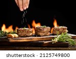 Cooked juicy steak meat beef with hand sprinking seasoning on top on wooden chopping board with flames in the background.