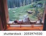 Relaxing Woman wrapped white towel lying on the wooden bench in Hot Finnish sauna with a huge wide window with green forest view and enjoying pleasant healthy body care temperature treatment.