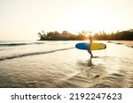 Teen boy with blue and yellow surfboard entering the waves for surfing with sunset rays on palm trees grove. Happy childhood and active sporty people vacation time concept.