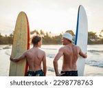 Small photo of Father with teenager son standing with surfboards on the sandy ocean beach with palm trees on background lightened with sunset sun. They smiling and have a conversation. Family active vacation concept