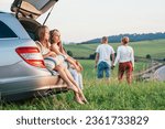 Two sisters girls sitting in car trunk enjoying sunset light. Young caucasian couple have auto trip break with hand in hand walk by green grass meadow. Family values, traveling and friendship concept.