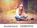 Young fashionable teenage girl with smartphone, camera and takeaway coffee in park in autumn sitting at smiling. Trendy young woman in fall in park texting. Retouched, vibrant colors.