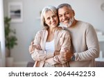 Small photo of Close up of beautiful smiling senior family couple husband and wife looking at camera with tenderness and love while standing in living room at home, retired man and woman embracing indoors