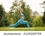 Small photo of Mental and physical health. Slender focused girl in sportswear performing yoga outside in early morning in nature, female standing in warrior pose with hands raised on grass in city park