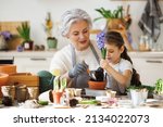 Cheerful granny and smiling granddaughter transplanting spring flower in pot while sitting together at table in kitchen