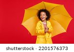 Small photo of Cheerful african american boy in raincoat laughing and carrying yellow umbrella on shoulder on rainy day against red background