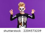 Small photo of Happy cheerful boy in skeleton costume celebrates Halloween and scary gesture on bright purple background