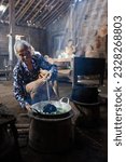 Small photo of Yogyakarta, Indonesia - Feb 12 2023: an old woman cooking on charcoal clay stove smiling in rustic house setting, rather noised and dark mood photo
