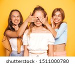 Three young beautiful smiling hipster girls in trendy summer clothes.Sexy carefree women posing near yellow wall in studio.Models surprising their friend.They cover her eyes and hugging from behind