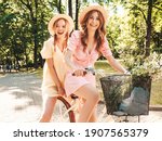 Two young beautiful smiling hipster woman in trendy summer sundress. Carefree women riding retro bicycle. Positive models having fun on bike posing in the park in hats. Best friends outdoors