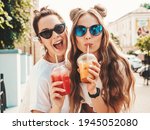 Two young beautiful smiling hipster female in trendy summer clothes. Carefree women posing outdoors.Positive models holding and drinking fresh cocktail smoothie drink in plastic cup with straw