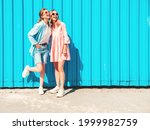 Two young beautiful smiling hipster female in trendy summer clothes and dress.Sexy carefree women posing in the street near blue wall.Positive pure models having fun at sunset, hugging and going crazy