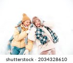 Two young beautiful smiling hipster female in trendy warm clothes and scarfs.Carefree women posing in the street in park. Positive pure models lying in snow. Enjoying winter moments. Xmas concept