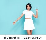 Young beautiful smiling female in trendy summer white top and skirt. Carefree woman posing near blue wall in studio. Positive model having fun indoors. Cheerful and happy. Celebrating and dancing 