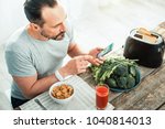 Small photo of Is under control. Concentrated careful unshaken man sitting in the kitchen by the table using his cellphone preparing for eating.