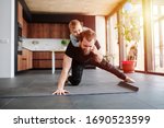 Father working out, doing single arm plank with his jolly infant baby riding on his neck. Stay at home apartment. Family quarantine, domestic life in self-isolation. Sunset light 