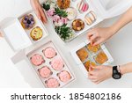 Female hands beautifully and carefully arranging colorful sweets in white windowed boxes. Zefir, cookies, macarons and cakes decorated with branches and flowers. Top view.