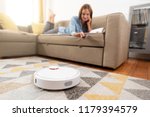 Robotic vacuum cleaner cleaning the room while woman relaxing on sofa. Woman controlling vacuum with remote control.