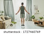 Small photo of Back view of young man in activewear jumping with skipping-rope while standing in front of window of living-room during workout