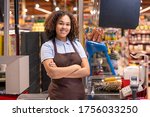 Small photo of Pretty female in apron standing by cashbox in supermarket and crossing arms by chest on background of shelves with food products