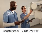 Small photo of Two young interracial clincians in uniform switching on medical equipment while female pressing button on tableau