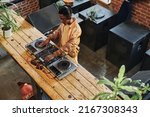 Small photo of Overview of young African American male deejay standing by long wooden table with dj set and creating new music