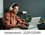 Small photo of Young smiling man in headphones typing on laptop keyboard while sitting by workplace and taking part in online webinar or lesson