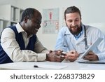 Small photo of Young smiling doctor pointing at tablet screen with results of medical test and explaining diagnosis to senior African American male patient