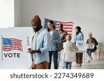 Small photo of Group of young multicultural voters in casualwear standing in queue along vote booths in polling place and putting their ballots into boxes