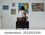 Rear view of young woman sitting on couch and looking at paintings on the wall, she enjoying art in gallery