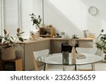 Part of spacious kitchen with round glass table with vases and notepads standing in the center against green domestic plants and counter
