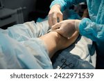 Small photo of Gloved hands of professional surgeon holding syringe with anaesthesia injection by knee of young female patient on operation table