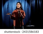 Small photo of Young comedian in eyeglasses and brown shirt standing on stage against blue curtains and speaking in microphone during performance