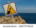Close-up of a danger sign for people falling down a ravine, crooked. In the background the Mediterranean Sea on the island of Mallorca at sunset