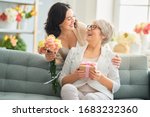 Happy mother's day! Beautiful young woman and her mother with flowers and gift box at home.                                