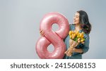 Small photo of Beautiful young woman with yellow flowers and a balloon in the shape of an eight on gray wall background.