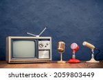 Small photo of Retro outdated analog CRT television and old microphones on wooden table front textured concrete black wall background. Vintage style filtered photo