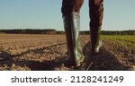 Small photo of Low angle: man walking in rubber boots in a farmer's field, the blue sky above the horizon. Man walking through an agricultural field. Farmer walks through a plowed field in early spring.