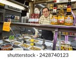 Small photo of Barcelona, Spain – April 17, 2018: Horizontal portrait of the owner of an olives and canned food stall in the old Abaceria Central Market of the Gracia neighborhood