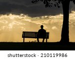 Silhouette portrait of a lonely anonymous man sitting on a bench watching the dark skies above roll by. 