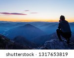 Male hiker crouching on top of the hill and enjoying scenic view of twilight landscape below. Hiking, achievement, expectation, optimism and self-reflection concepts.
